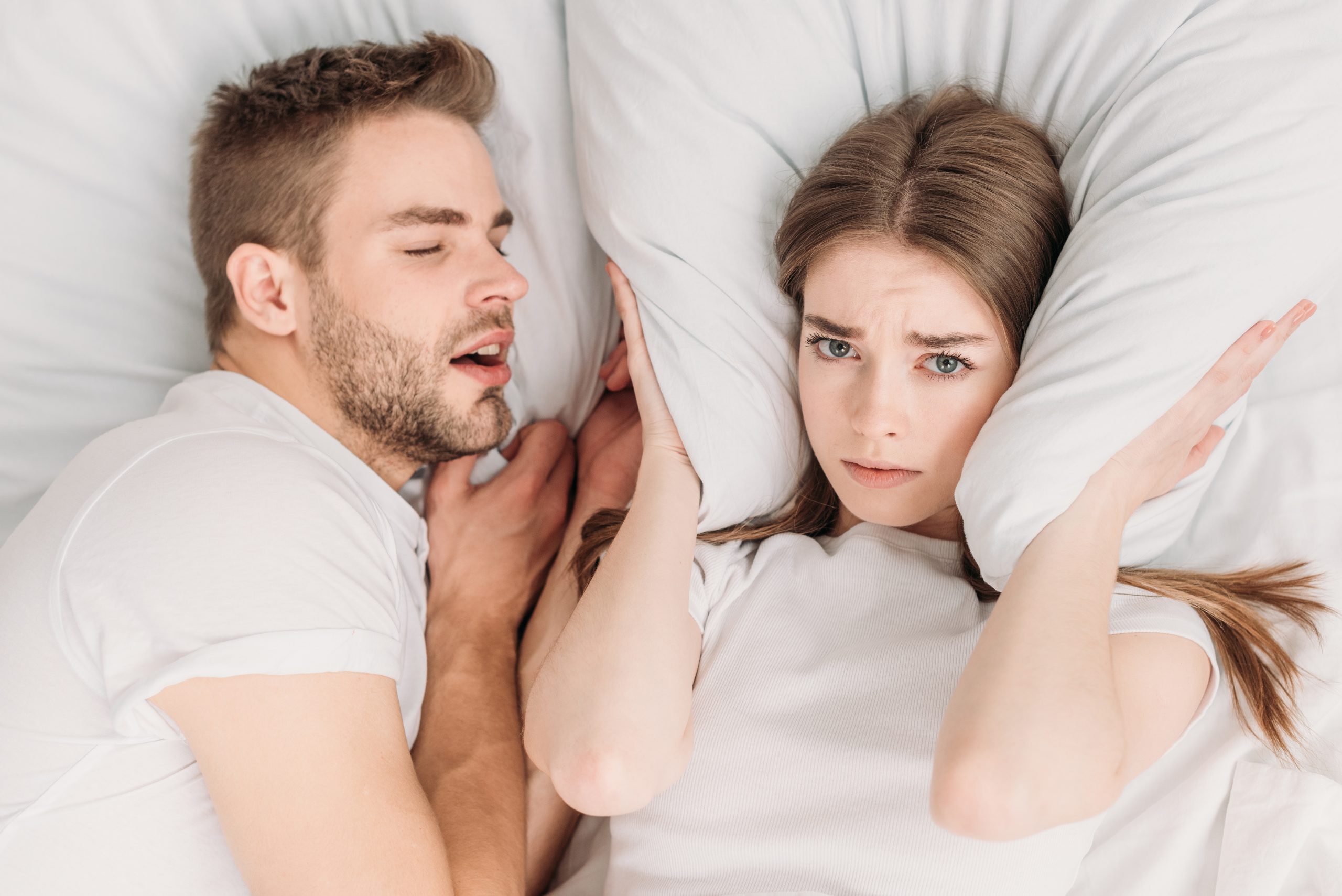 top view of woman plugging ears with pillow while lying in bed near snoring husband
