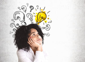 Smiling young African American woman in white shirt looking at question marks and lightbulb drawn on concrete wall. Concept of good idea. Mock up